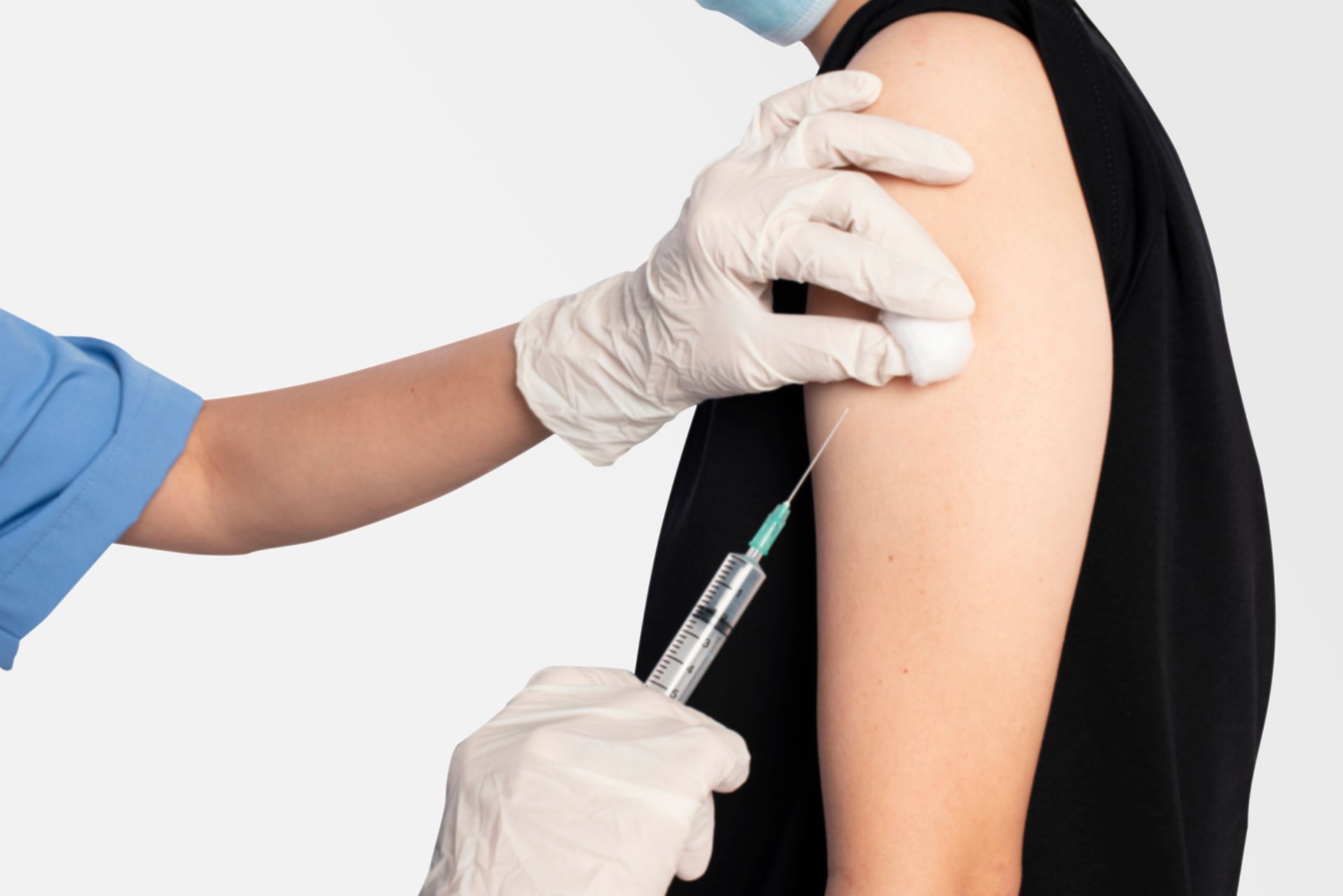 A person receives a vaccine injection in their upper arm. A gloved hand holds a cotton ball, another injects with a syringe against a white background.