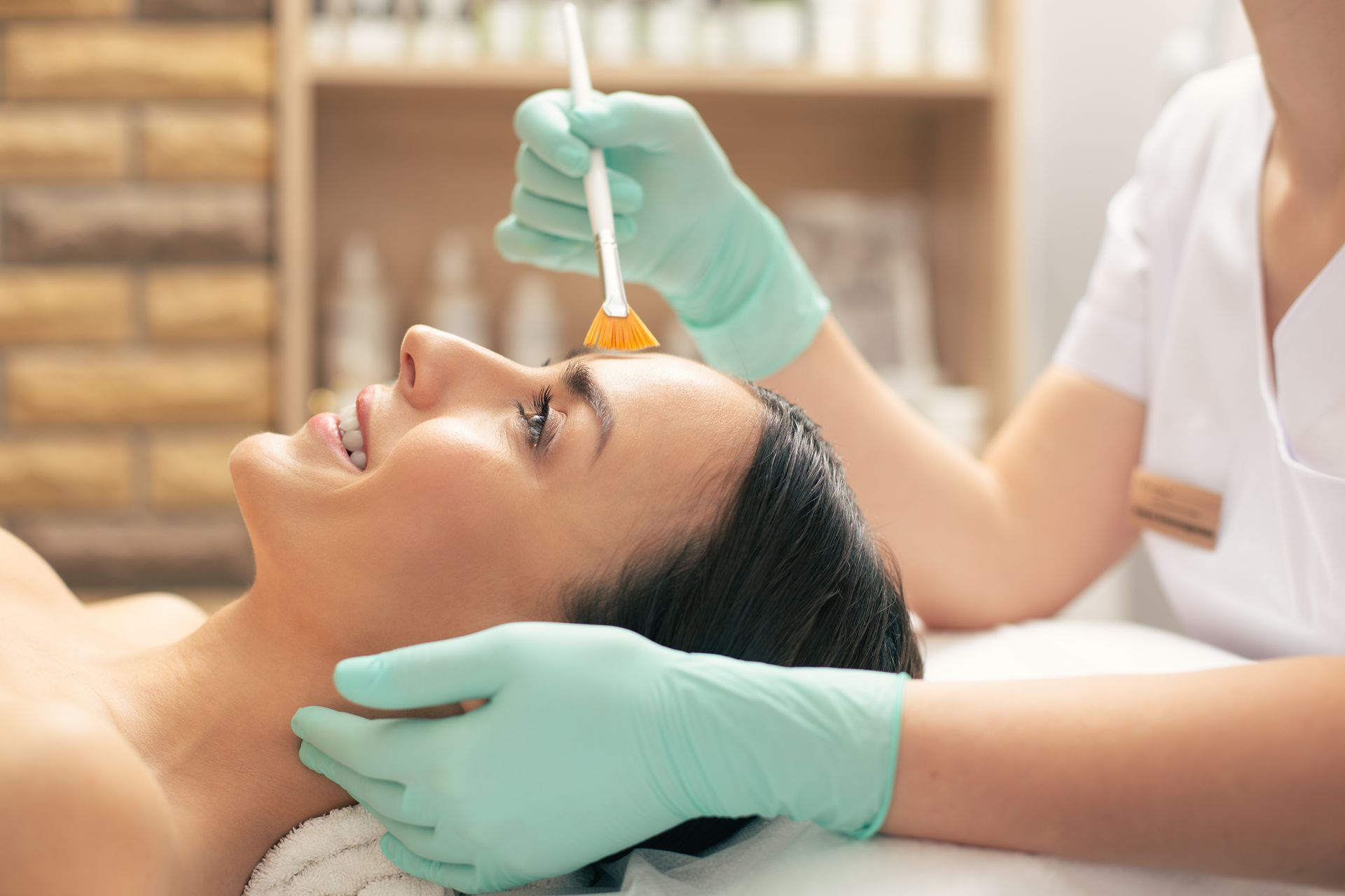 Woman receiving facial peel treatment, esthetician applying product with brush in spa.