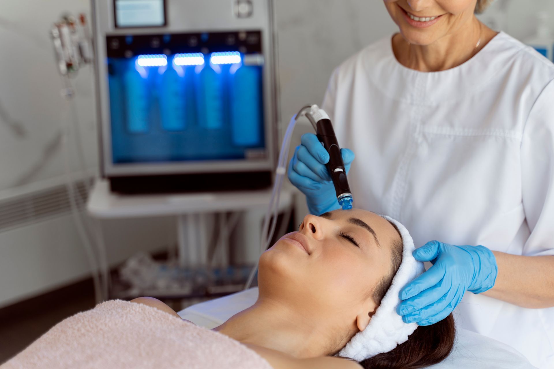 A practitioner in a white uniform and blue gloves performs a hydrafacial treatment on a client in a clinic.