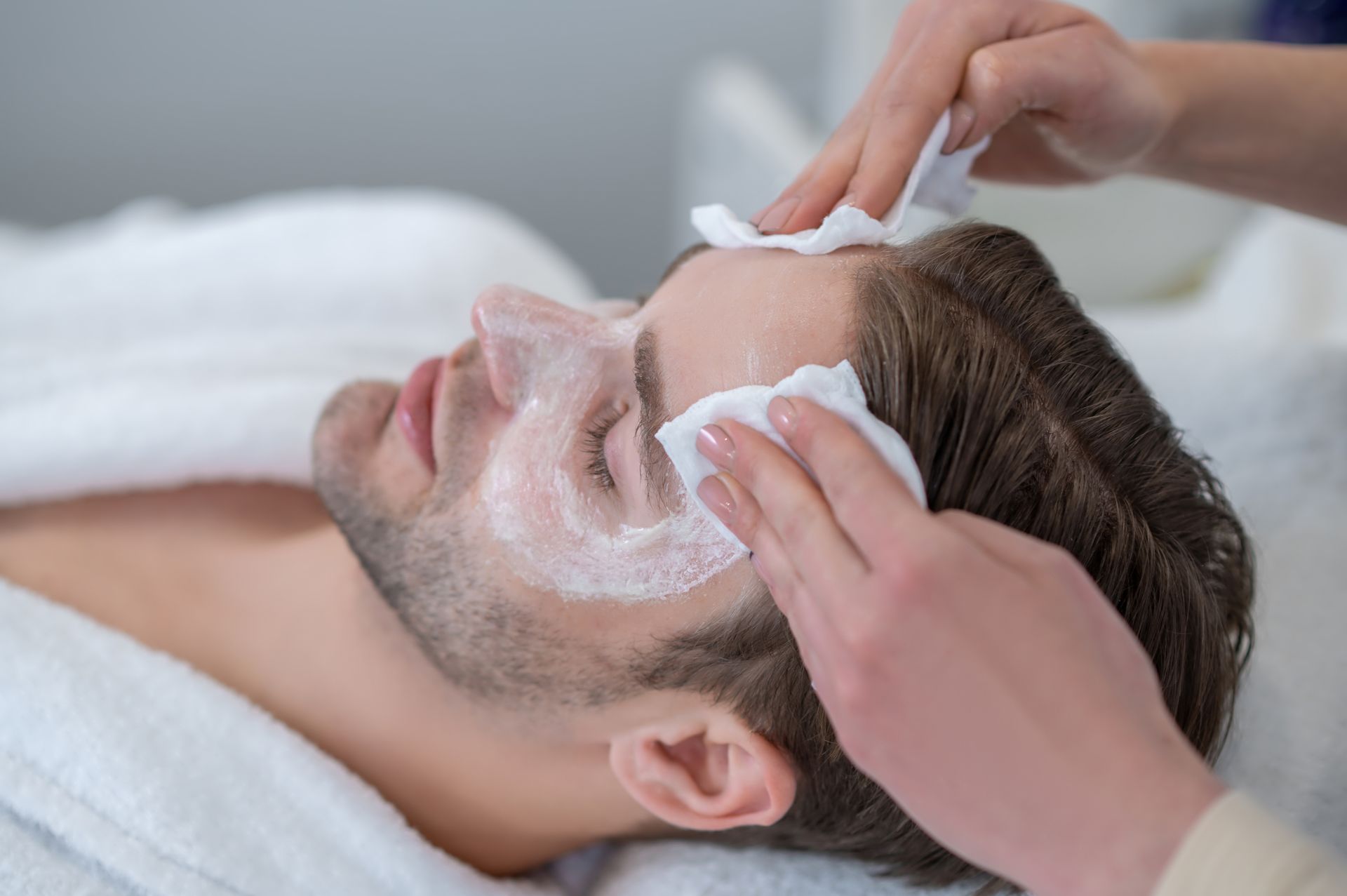 Man receiving facial treatment, with hands wiping his face with white cloths.