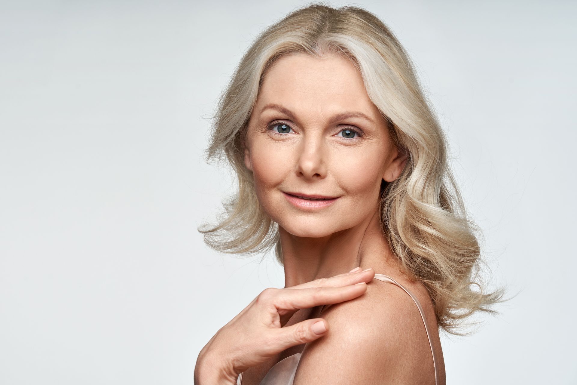 A person with shoulder-length blonde hair posing against a light background, resting one hand gently on their shoulder.