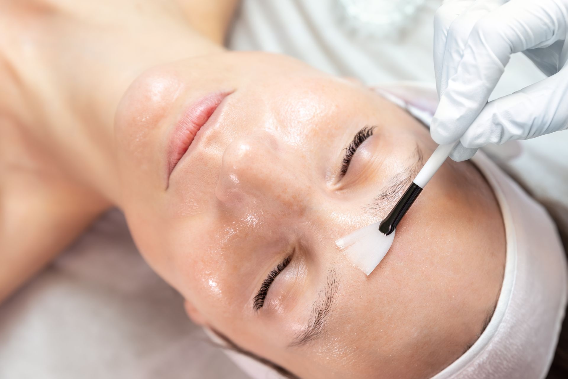 A person in a headband receives a professional facial treatment, with gloved hands using a fan brush to apply a solution.