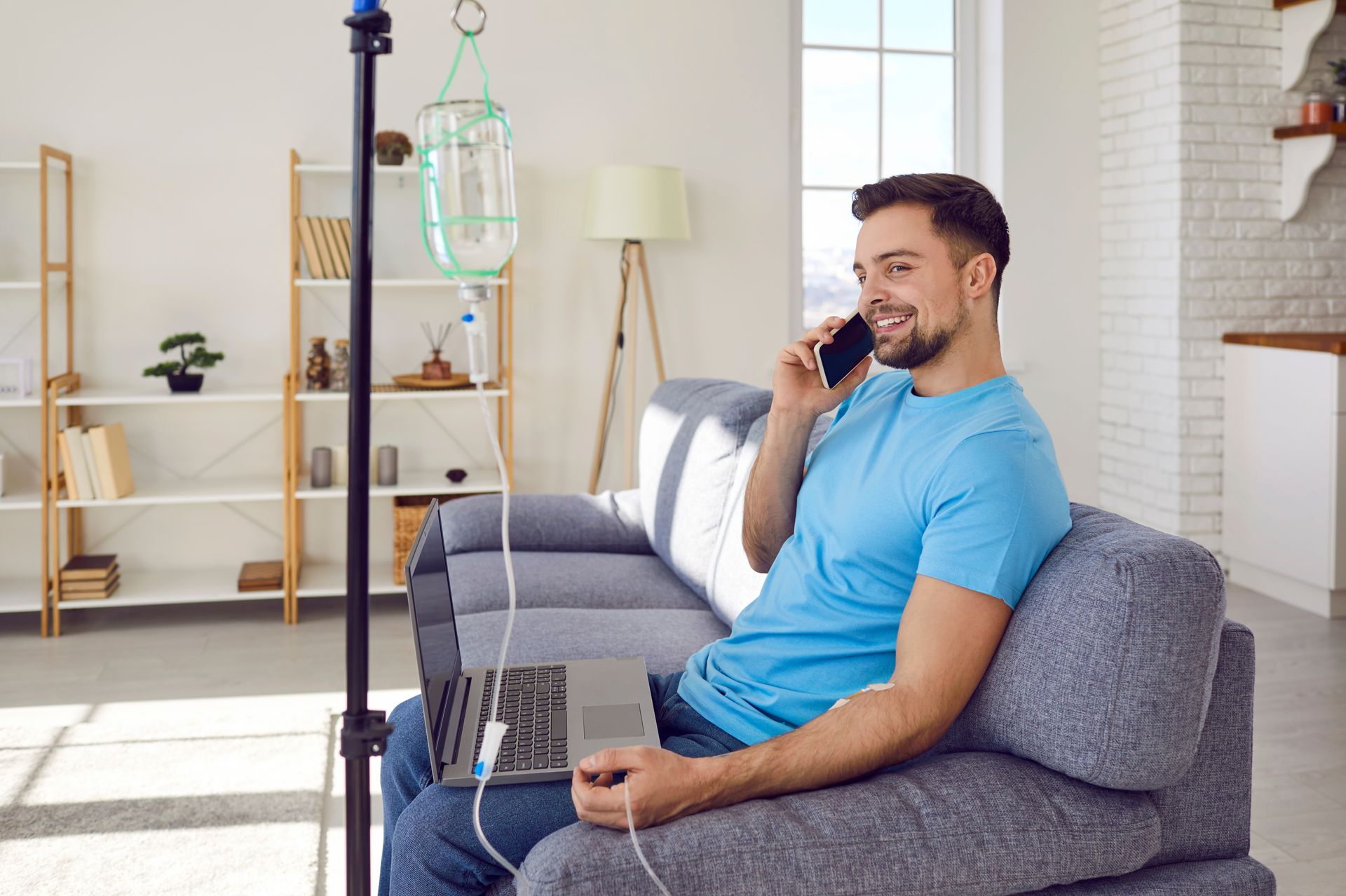 Man on couch with IV drip and laptop, talking on phone, in home setting.