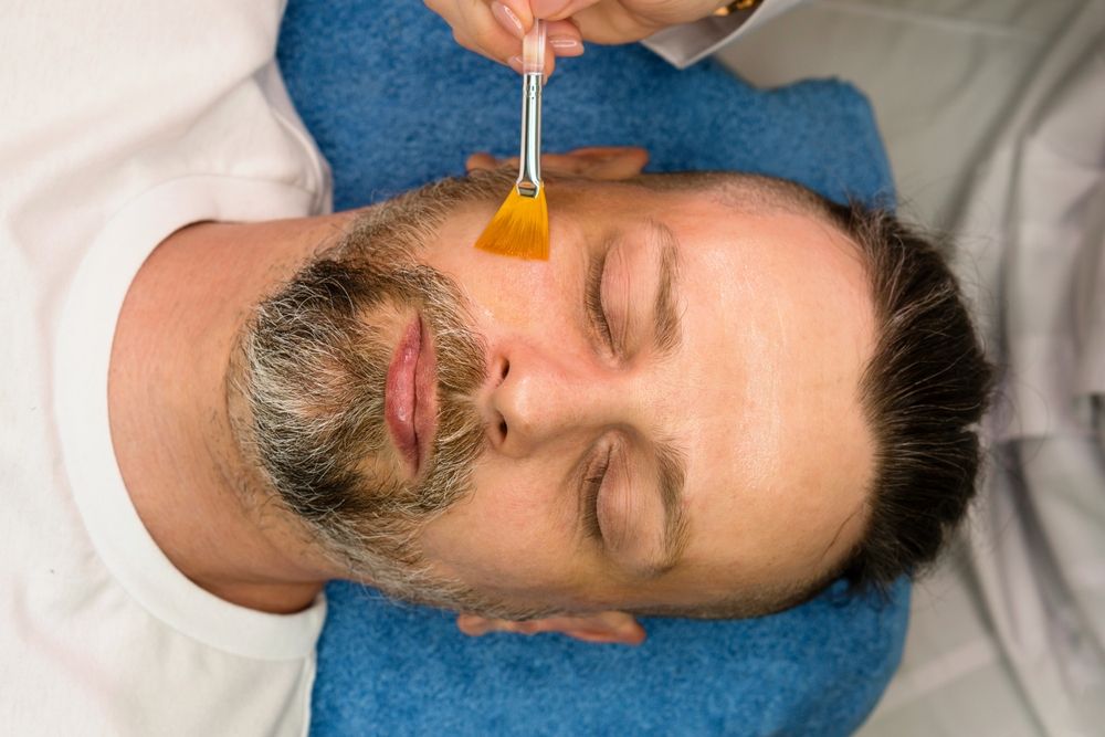 A person lying on a blue towel receives a facial treatment applied with a small yellow brush.