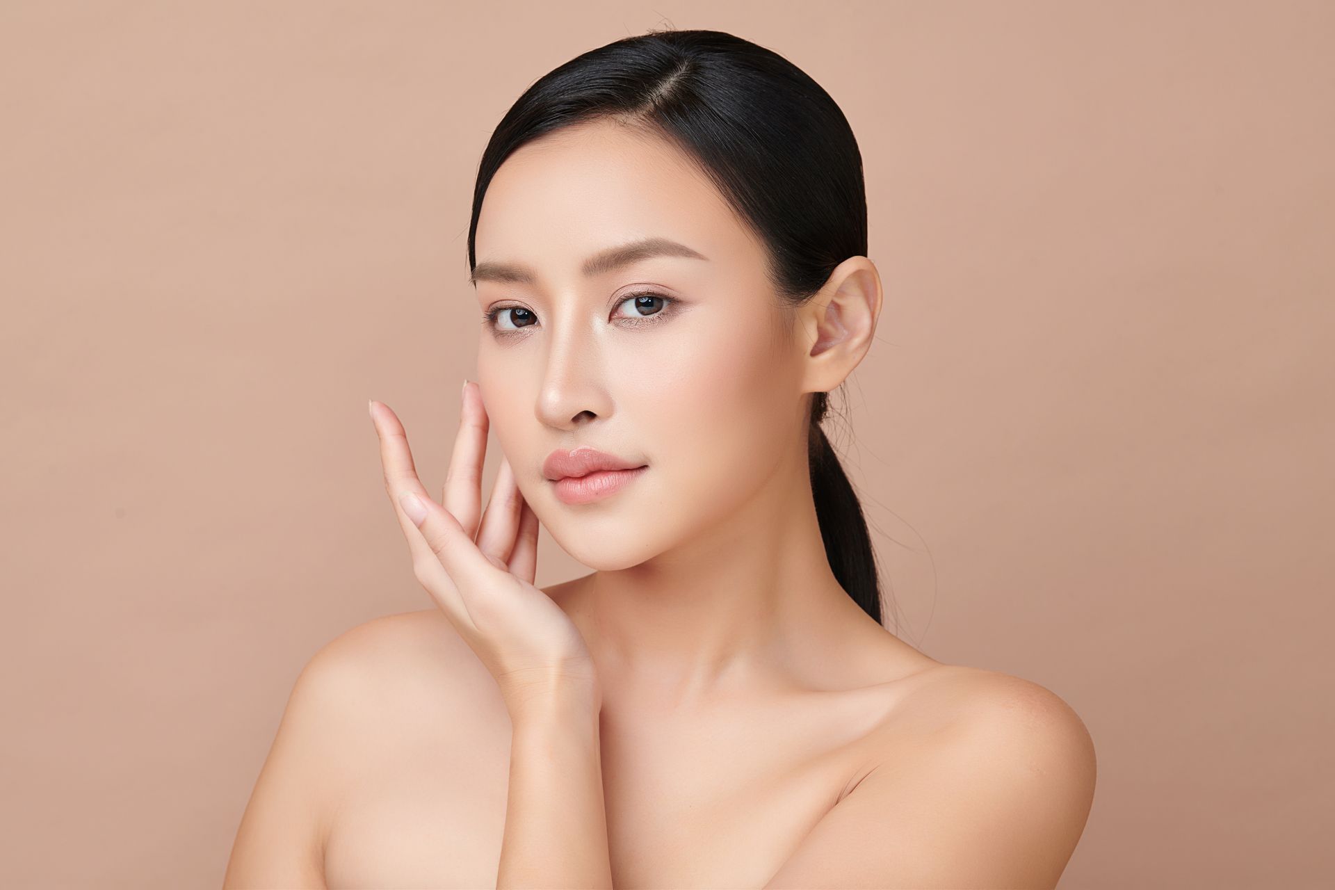 Woman with smooth skin, hand on cheek, looking at the camera against a neutral background.