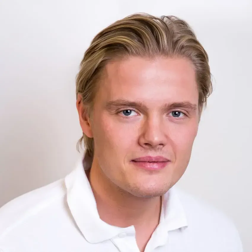 Blond mann med lyse øyne i hvit skjorte, smiler litt, foran en hvit bakgrunn.