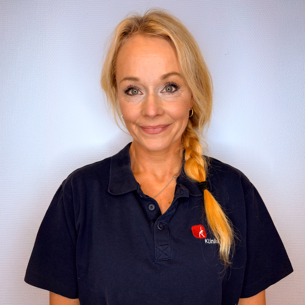 Kvinne med blondt hår i en lav hestehale, iført en mørkeblå poloskjorte, smilende foran en hvit vegg.