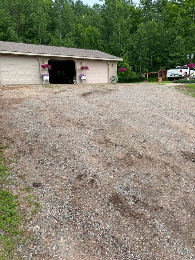 Gravel driveway leading to a garage in a wooded yard with people and vehicles nearby.
