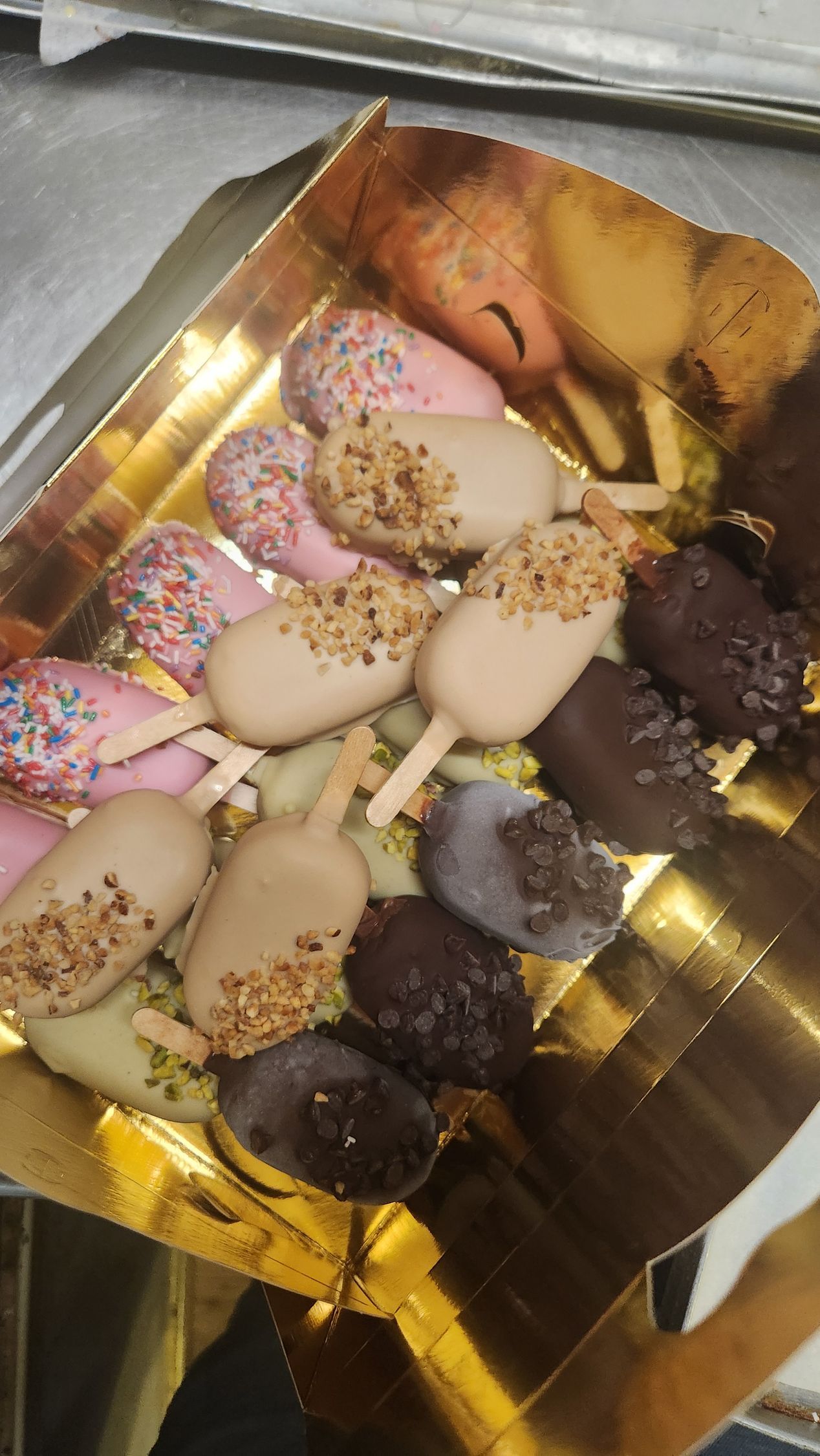 Cake pops assortiti ricoperti di cioccolato e decorati con zuccherini, disposti su un vassoio dorato.