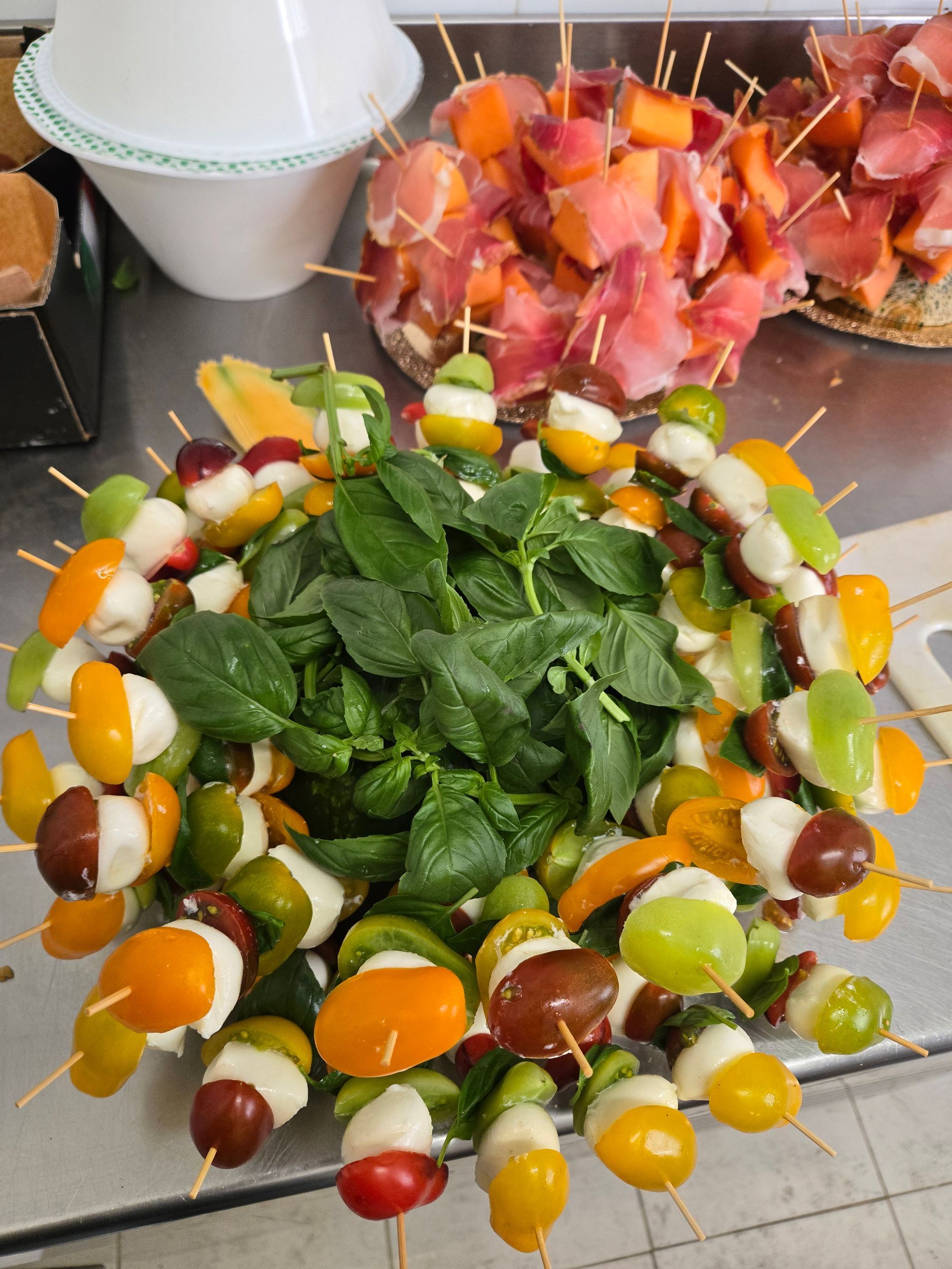 Spiedini di frutta disposti intorno a verdure a foglia verde su un vassoio, con bicchieri sullo sfondo.