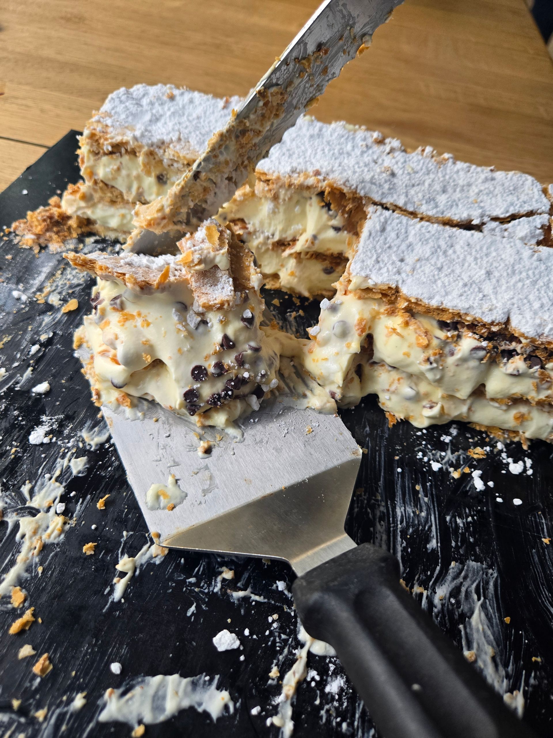 Una fetta di torta ricoperta di zucchero a velo con ripieno cremoso, disposta su un vassoio e tagliata con coltello e spatola.