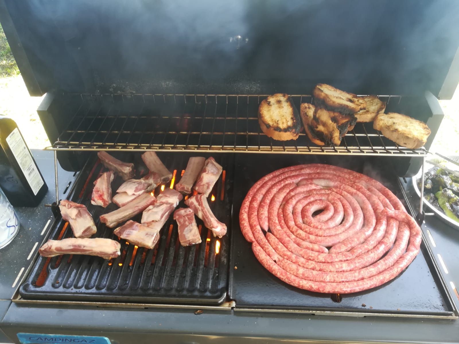 Griglia con salsicce, costine e pollo che cuociono su due livelli, con il fumo che sale.