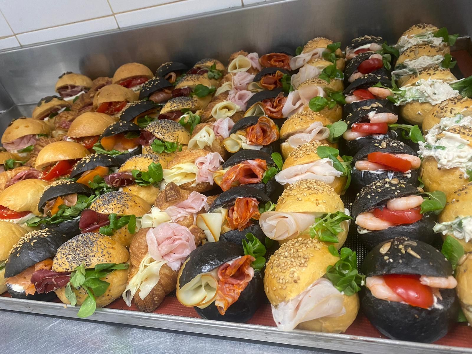 Assortimento di mini tramezzini ripieni disposti su un vassoio, guarniti con sesamo, verdure, pomodori e salse.