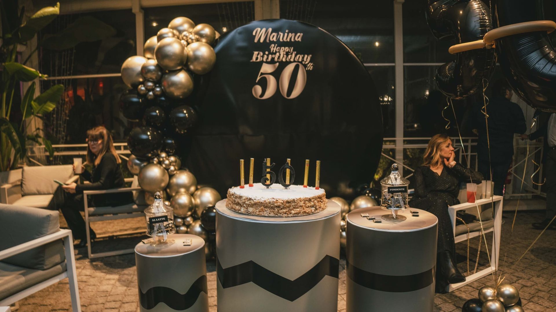 Allestimento per una festa di 50° compleanno con palloncini neri e oro, torta e tavoli da dessert decorati.