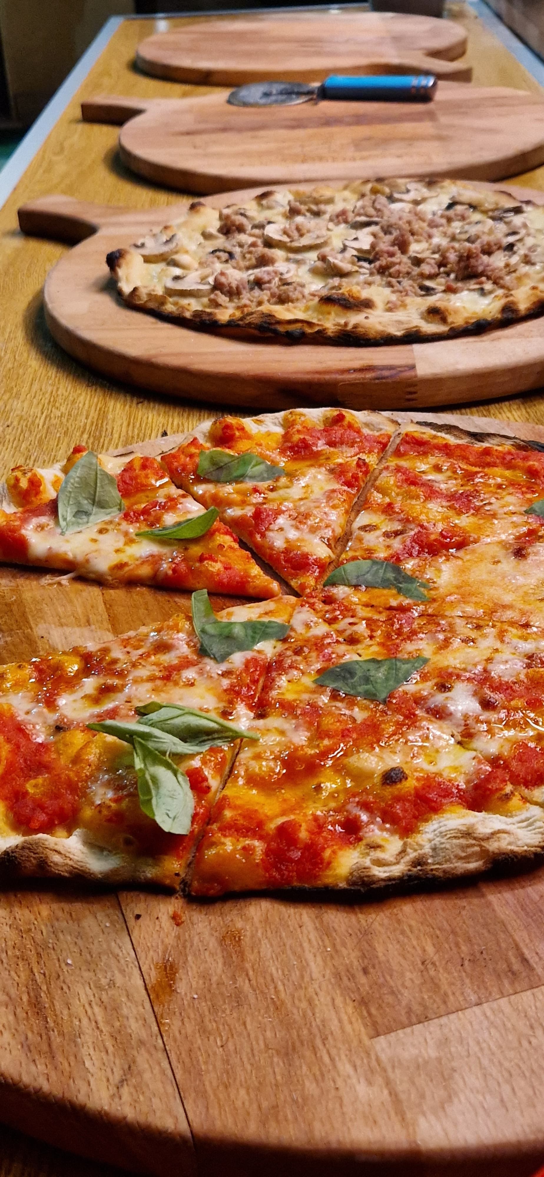 Vassoio di legno con tre pizze, tra cui una colorata pizza al pomodoro e basilico in primo piano.
