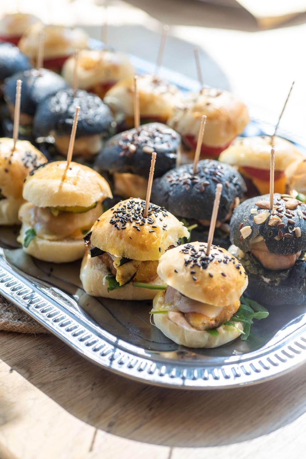Vassoio di mini-hamburger con panini al sesamo e semi di papavero, disposti con stuzzicadenti su un piatto d'argento