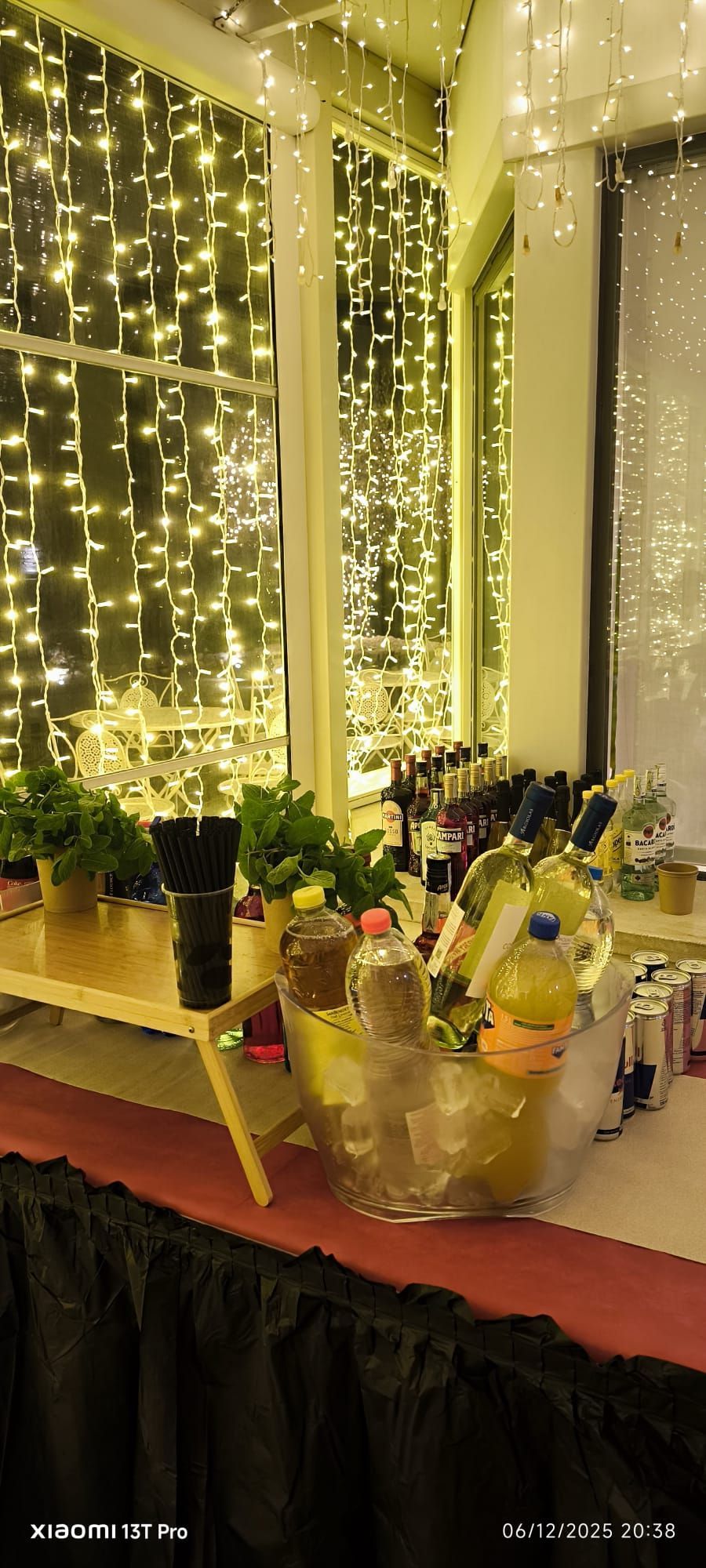 Tavolo festivo per dolci e bevande con fiori gialli, bottiglie e luci decorative sullo sfondo.