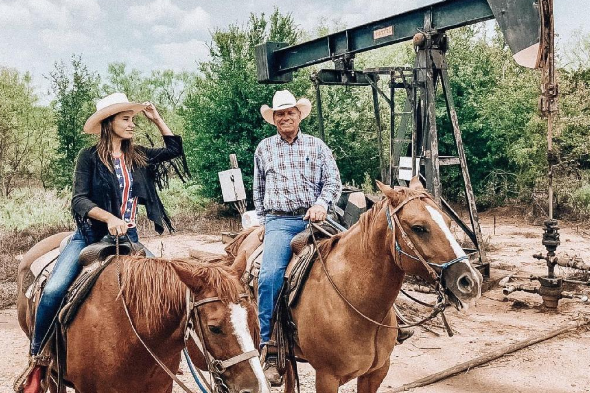 A man and a woman are riding horses in front of an oil pump.