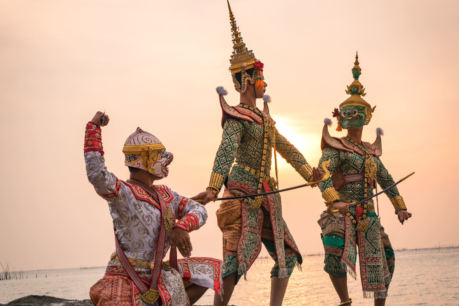 Three people in traditional costumes are dancing on the beach at sunset.
