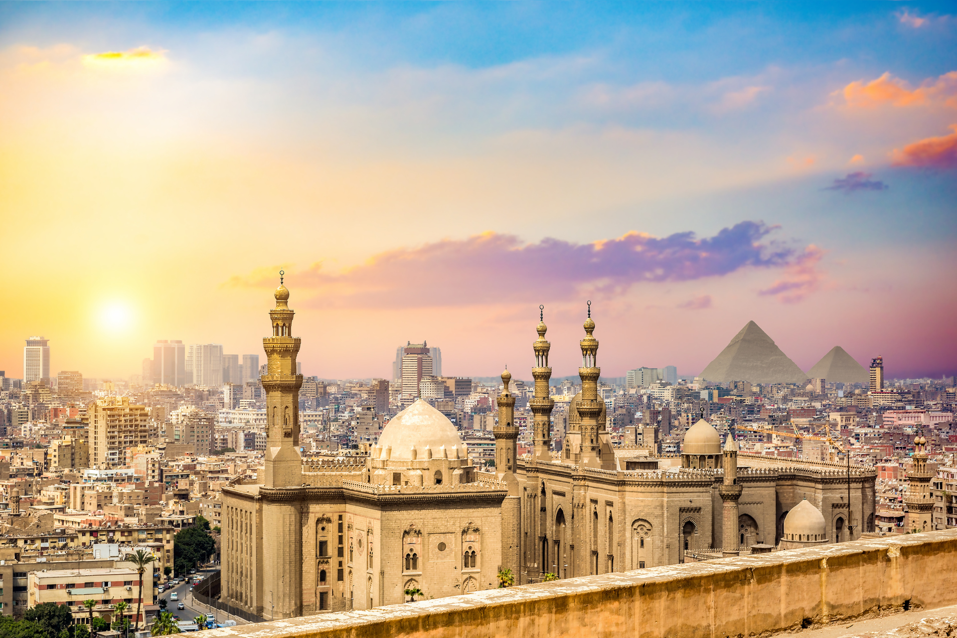 A cityscape with a mosque and pyramids in the background at sunset.