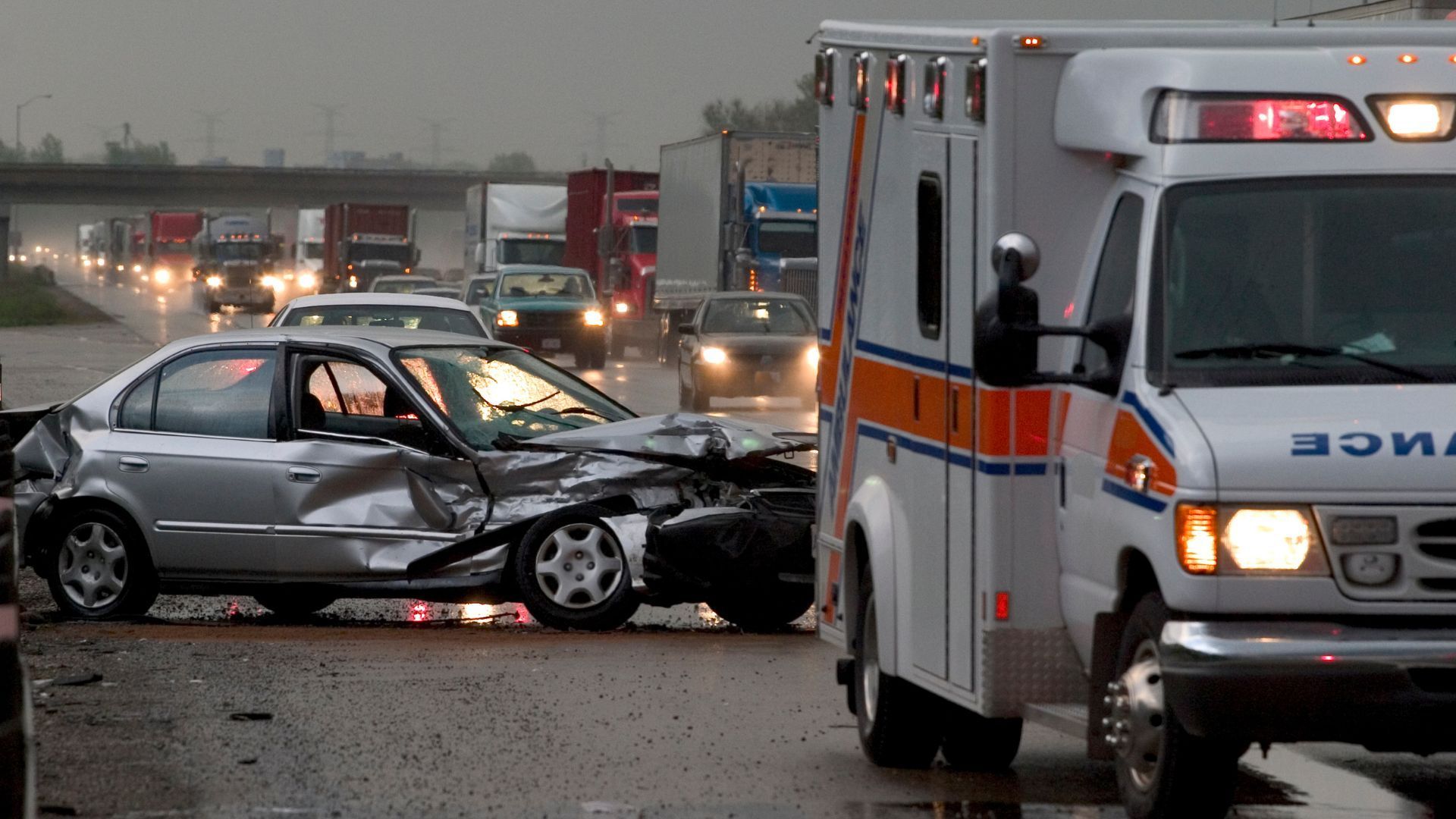 Damaged sedan after crash on a wet highway beside an ambulance and traffic emergency scene