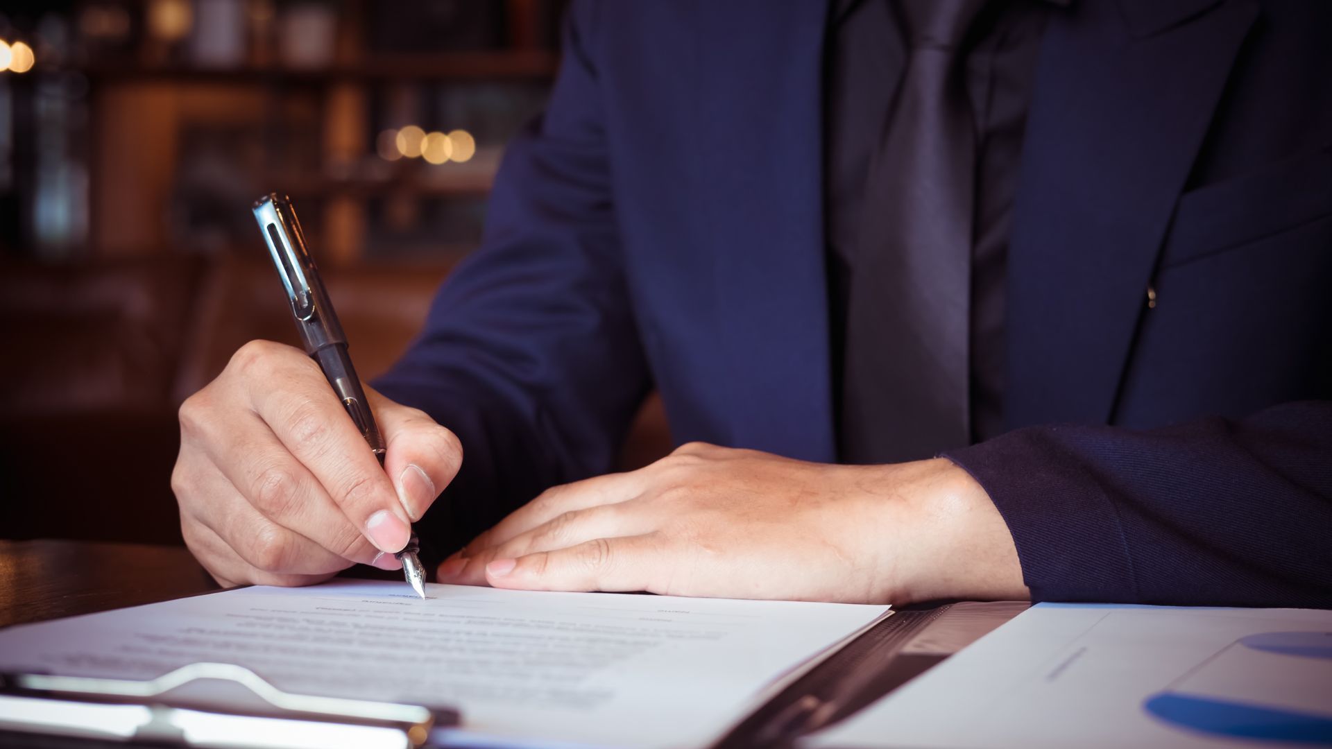 Person in a dark suit signing a document at a desk with a pen