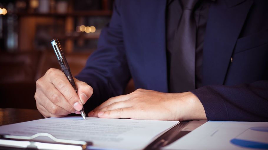 Person in a dark suit signing a document at a desk with a pen