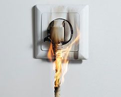 Burning electrical plug in a wall outlet, with flames rising from the cord