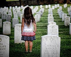 Young girl stands among rows of white gravestones in a cemetery, facing away.