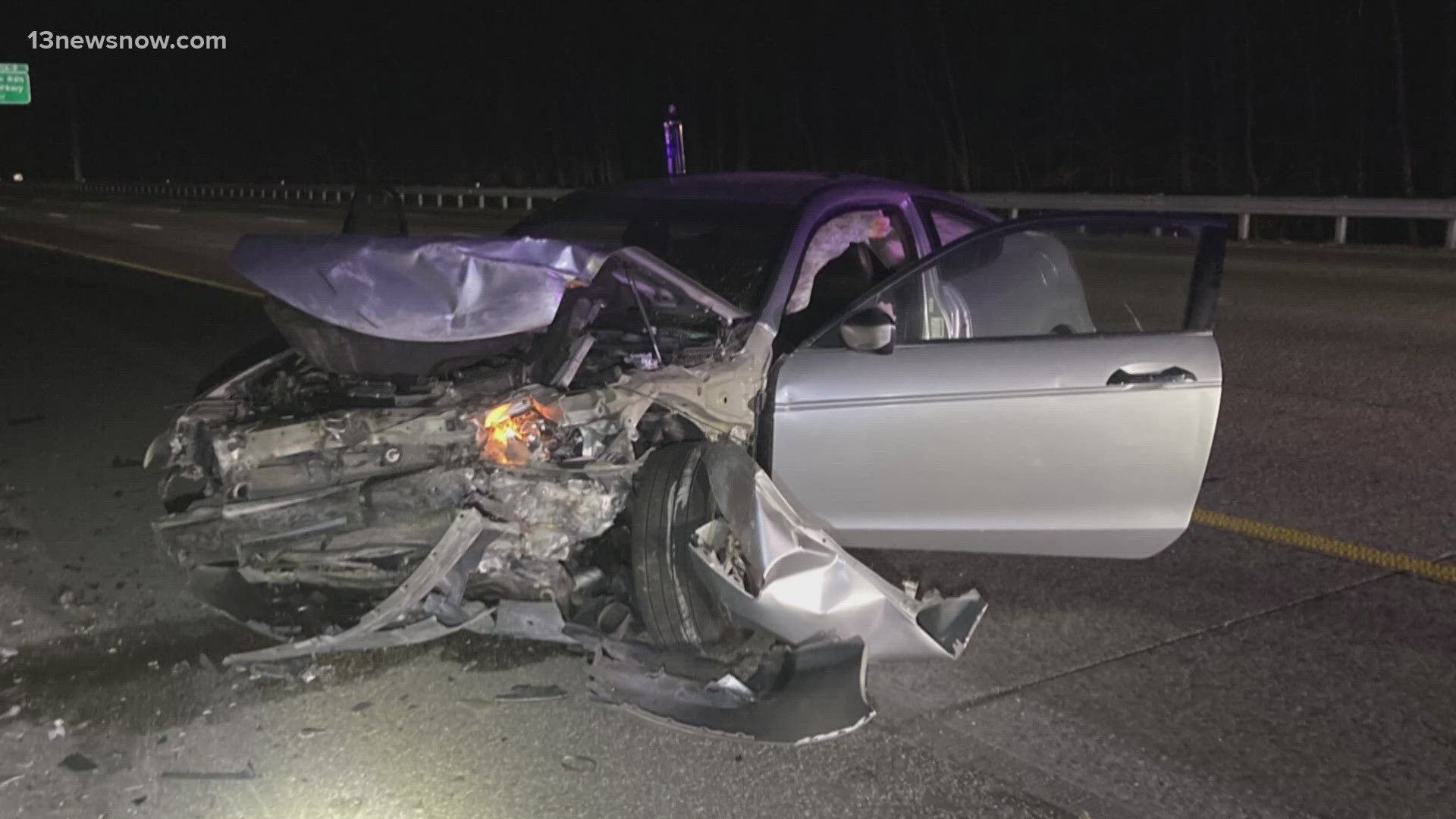 Severely damaged silver car at night on a road, with front-end wreckage and open passenger door