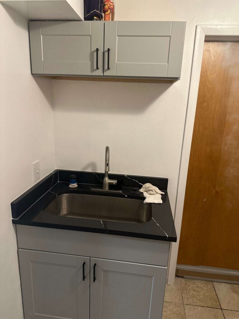 A utility sink area with gray cabinets, black countertop, stainless steel faucet, and a wall cabinet.
