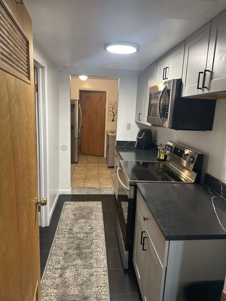 Narrow kitchen with gray cabinets, black countertops, and a long rug. Doorway to other rooms.