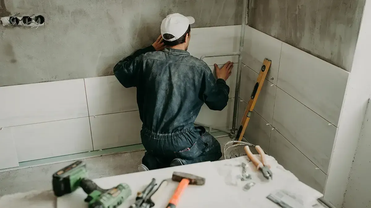 A person in blue coveralls and a cap installs tiles in a bathroom, with tools on a table.
