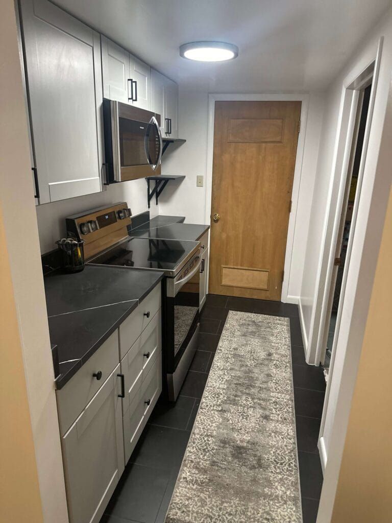 Narrow galley kitchen with gray cabinets, stainless steel appliances, black countertops, and a wood door at the end.