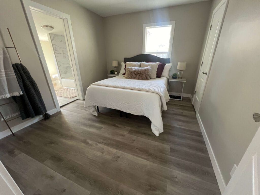 Bedroom with a bed, nightstands, a doorway to a bathroom, and wood-look flooring. Gray walls and white trim.