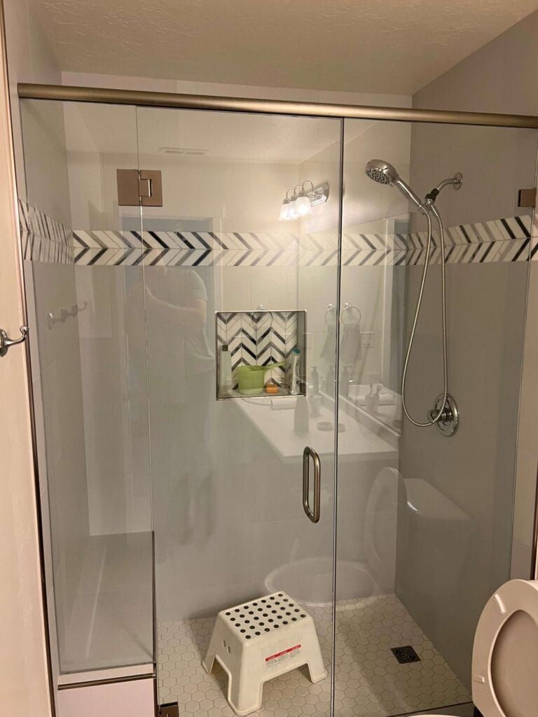 Glass shower enclosure with decorative tile band and small step stool inside.