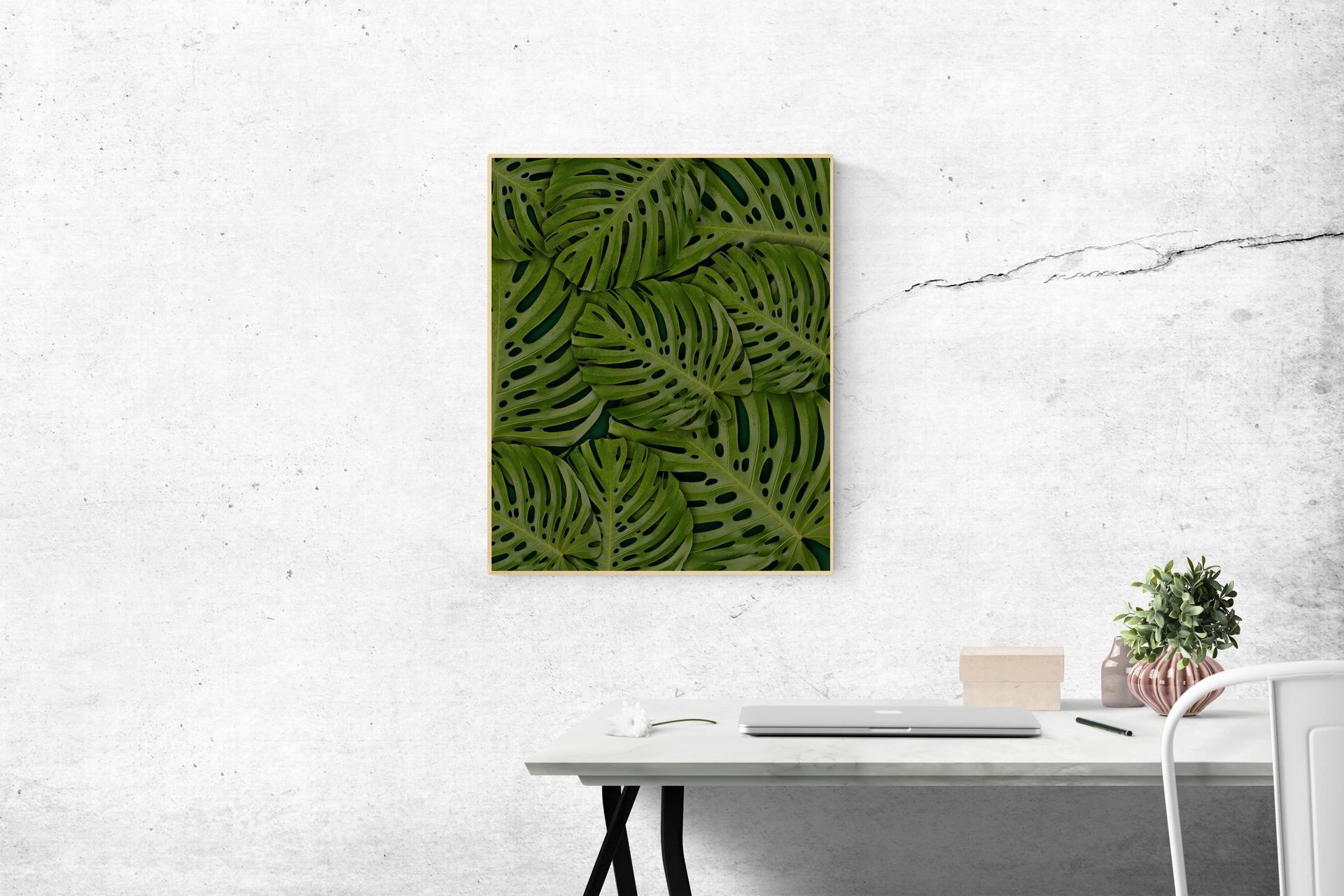 Canvas with green tropical leaves hangs on a white wall above a desk with laptop and plant.
