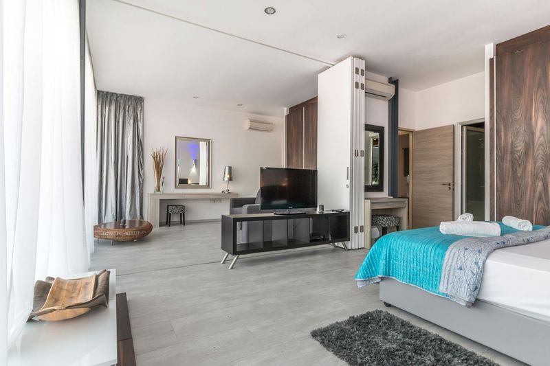 Modern bedroom with turquoise bedspread, TV, desk, and large windows with sheer curtains.