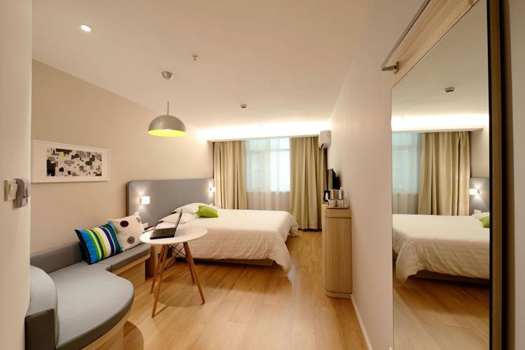 Hotel room with a bed, small sofa, table, and mirror. Beige walls, wood floor, and sunlight. Hotel room with a bed, small sofa, table, and mirror. Beige walls, wood floor, and sunlight.