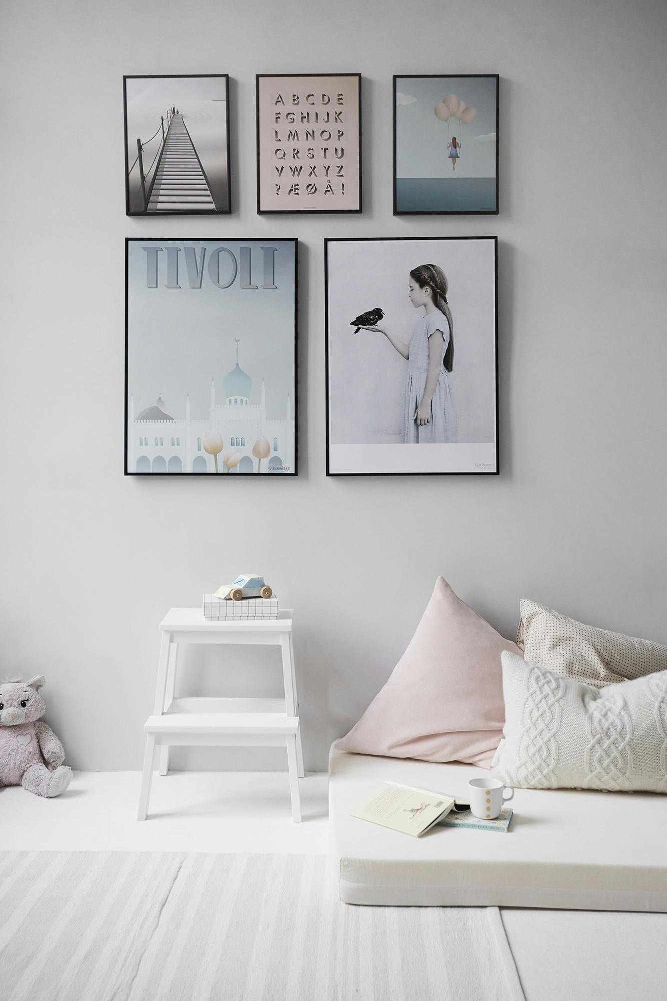 Framed pastel wall art above a white shelf and cushions in a soft, minimalist nursery.