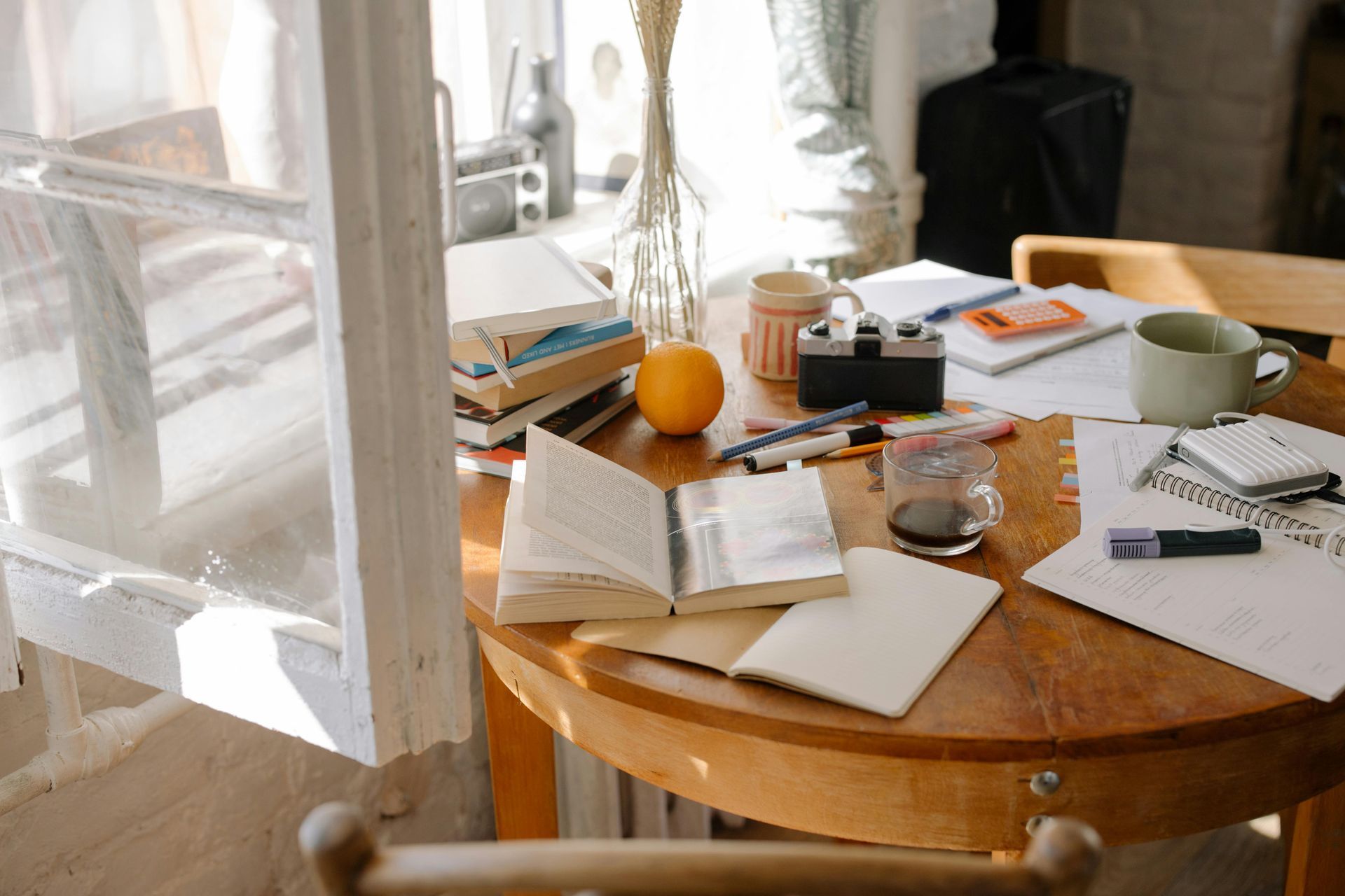 Round wooden table with books, camera, orange, and writing supplies near a window.