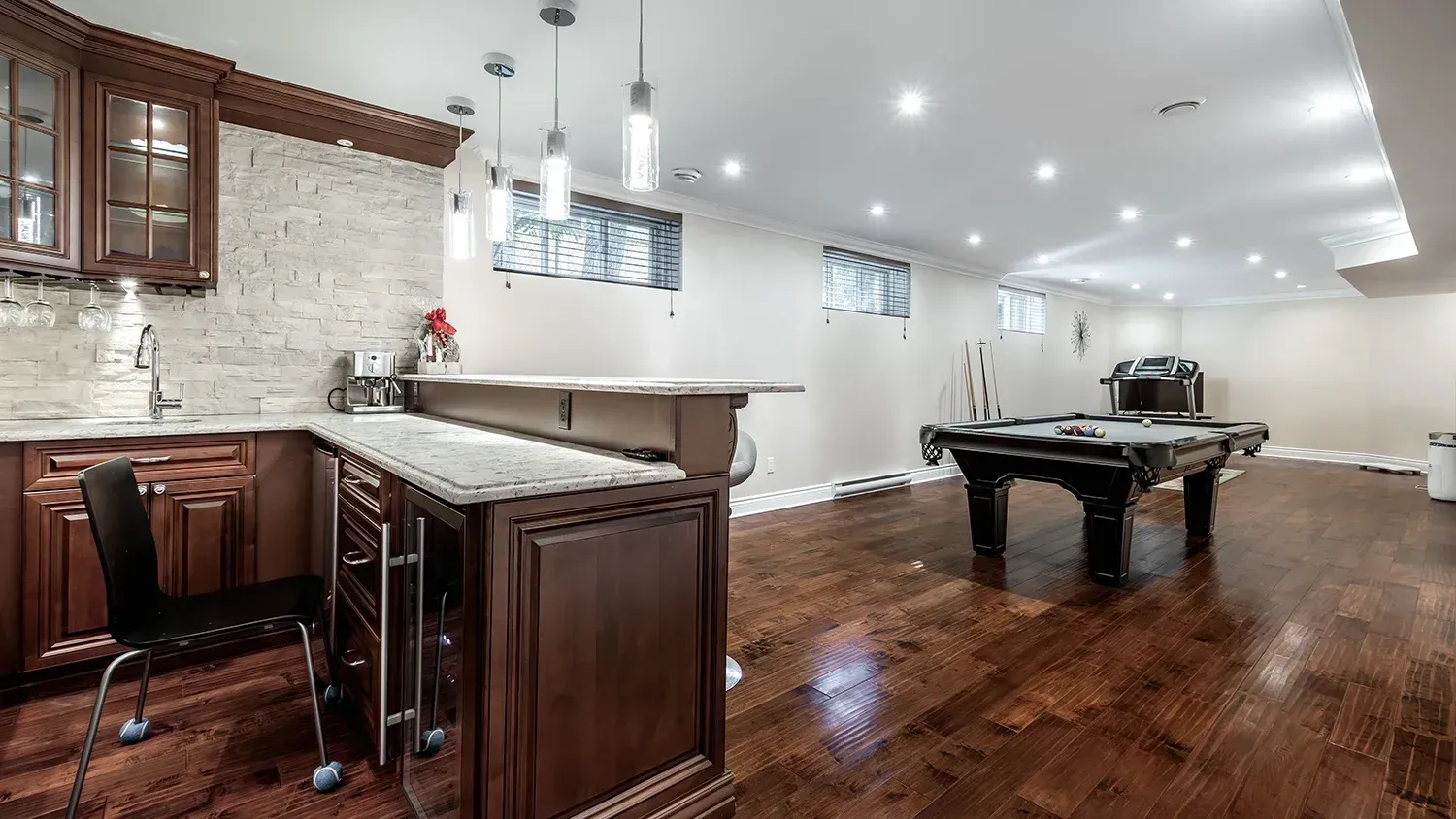 Basement with bar, kitchen, and pool table. Brown cabinets, light countertops, wood floor, white walls.