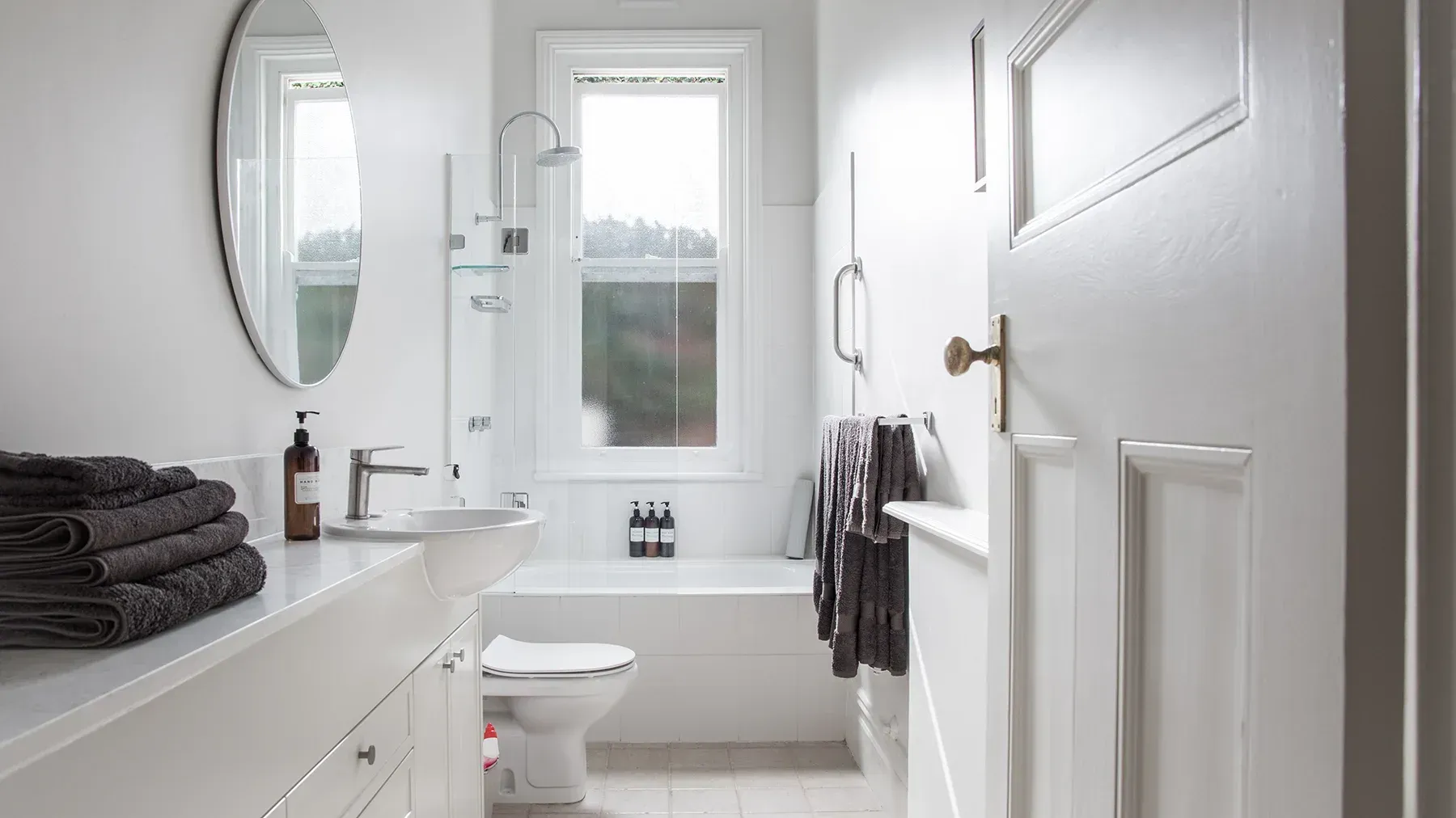White bathroom with a sink, toilet, shower, and towels. Open door on the right.