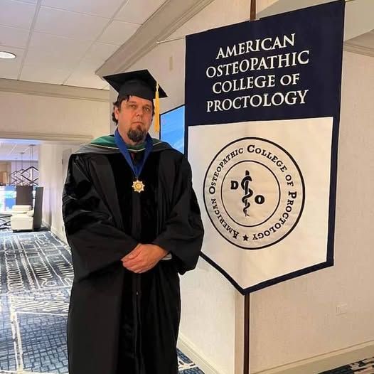 A man in a graduation cap and gown stands in front of an american osteopathic college of proctology banner