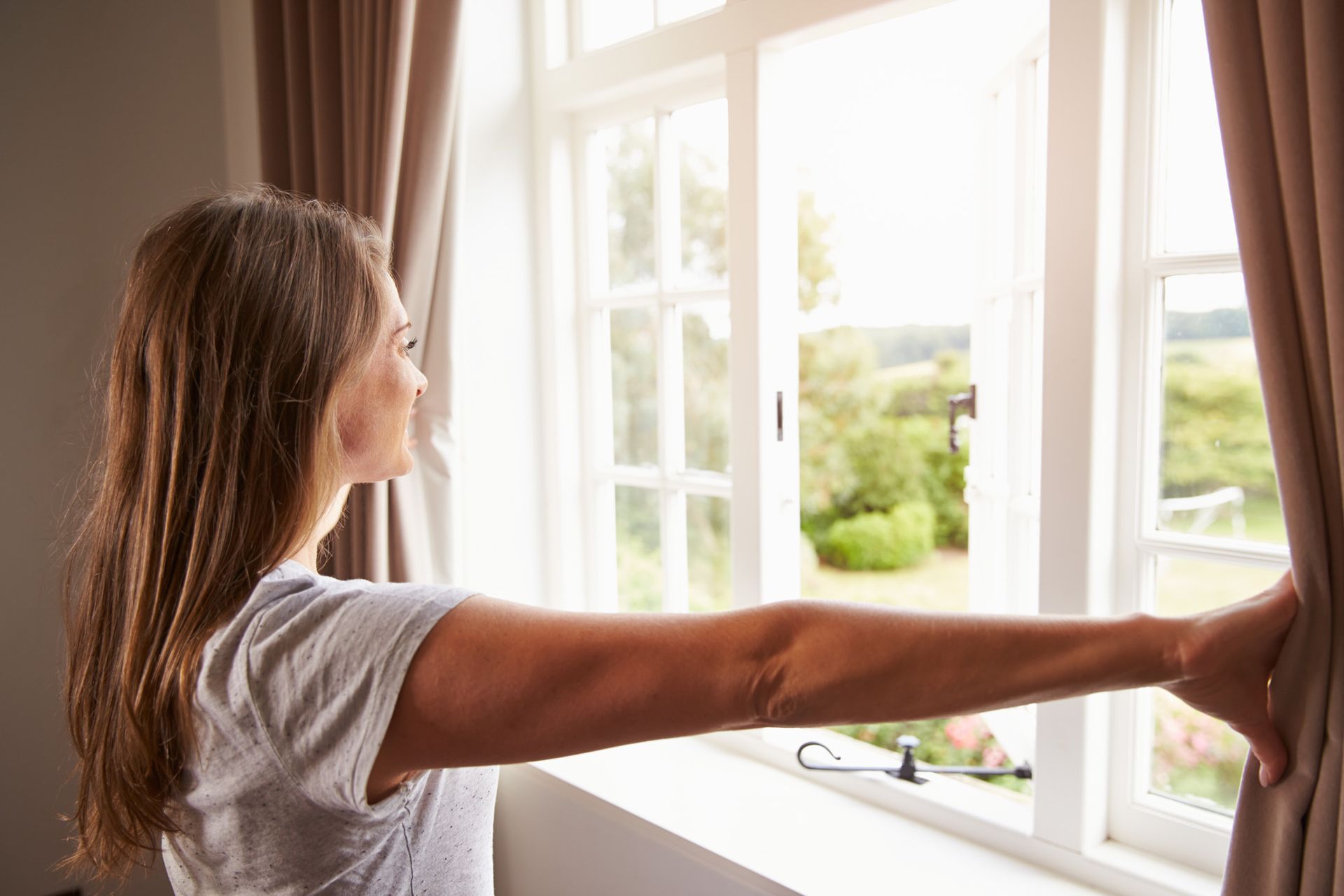 A woman is opening her curtains and looking out of a window.