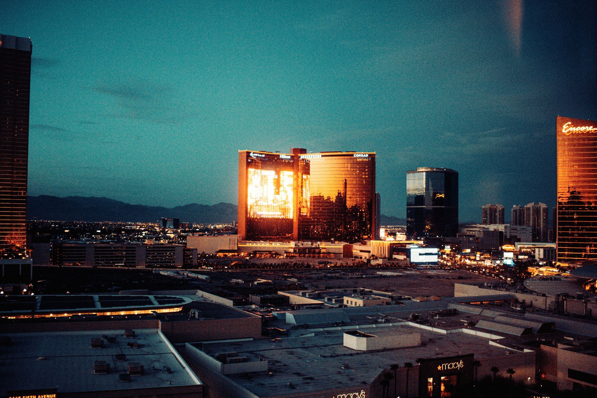 City skyline at dusk with a bright, reflective building glowing orange amid darkened high-rises and rooftops
