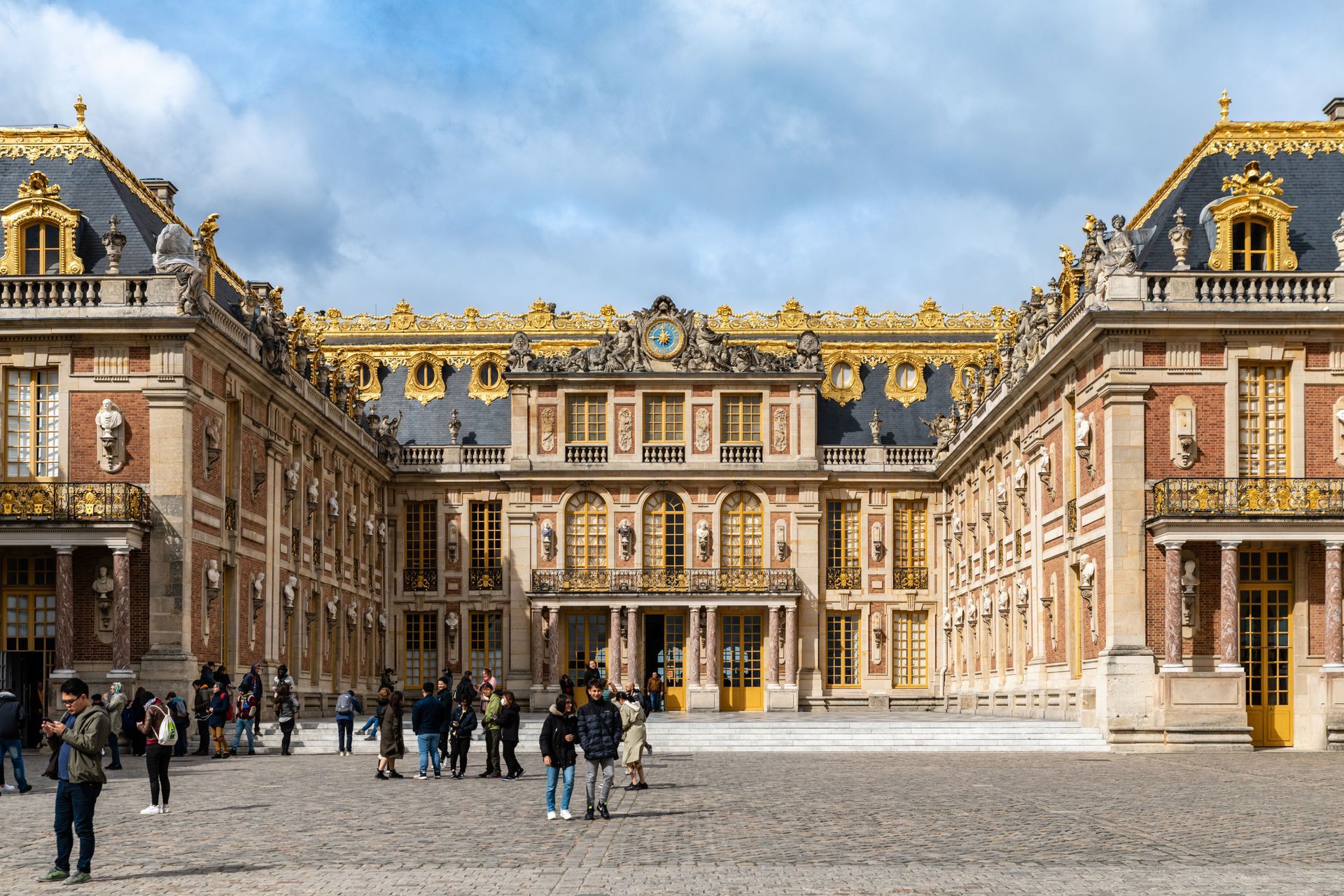 People in a courtyard before a grand ornate palace with gold-trimmed roofs under a blue sky