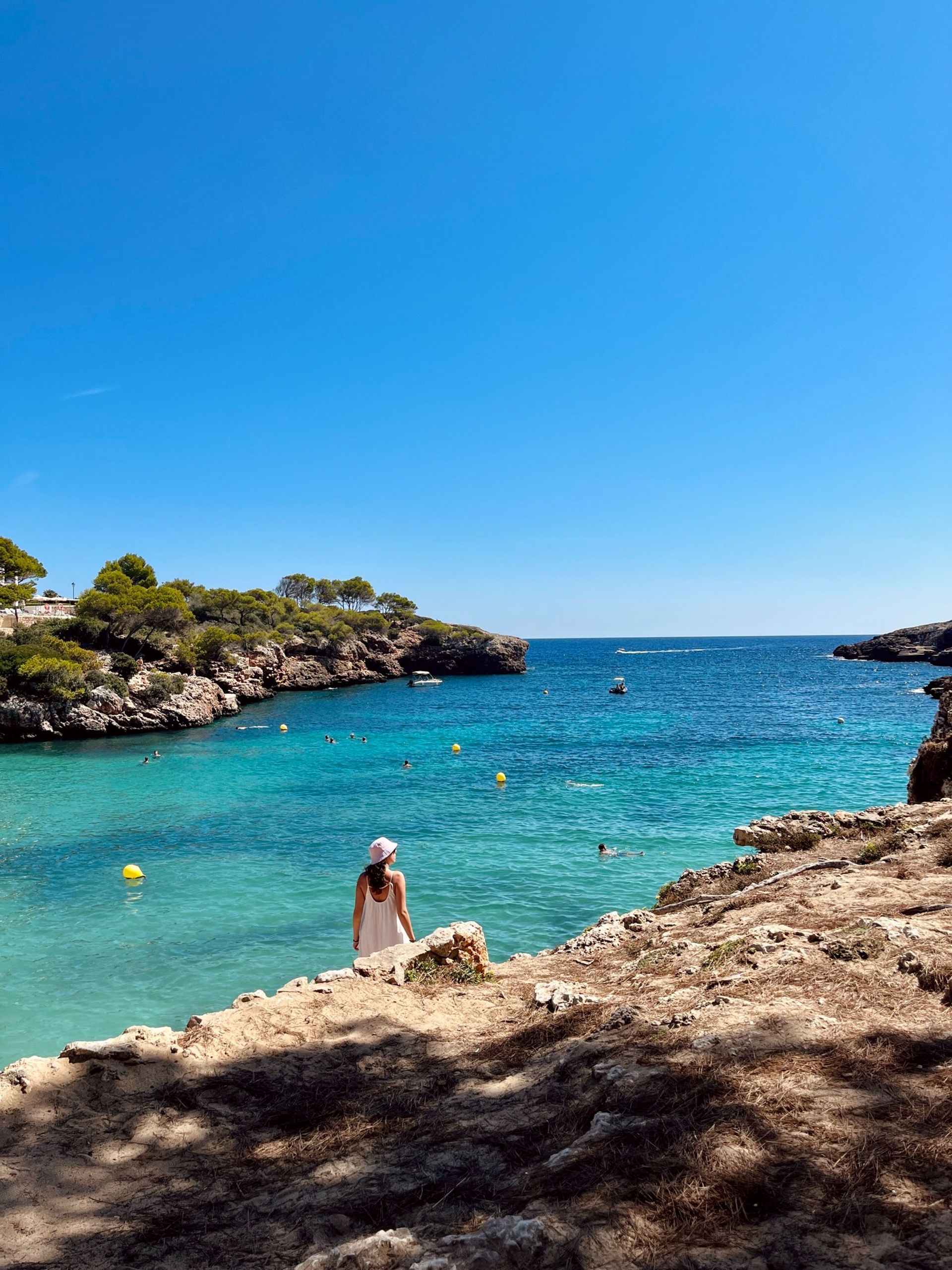 Person standing on rocky shore beside turquoise bay under a clear blue sky