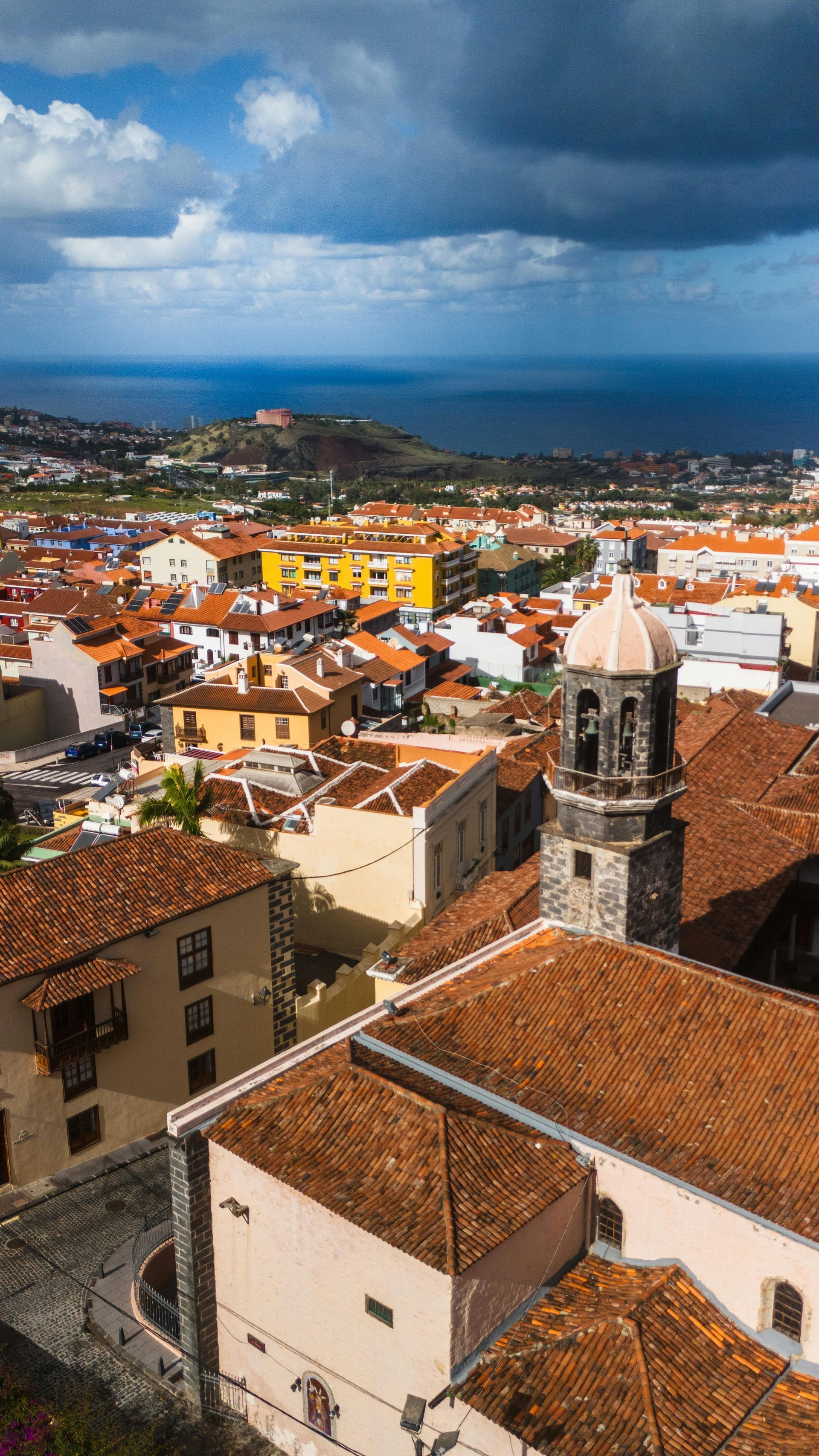 Historic hillside town with red-tiled roofs, church tower, and cloudy blue sky over a coastal city