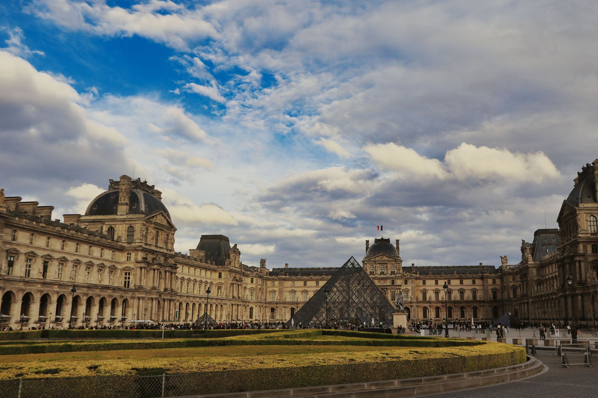 Courtyard of the Louvre Museum in Paris under a cloudy blue sky, with historic stone buildings and a glass pyramid